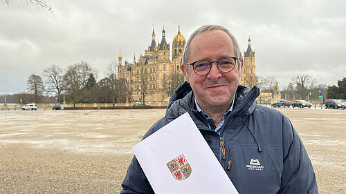 Es ist Prof. Baldauf mit seiner Urkunde in der Hand vor dem Schweriner Schloss zu sehen.