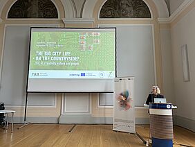 Conference kick-off at the Humboldt Graduate School. A woman speaks at a lectern in front of a large screen displaying the conference slide ‘The Big City Life on the Countryside? Yes, AI, creativity, nature and people’ in a historic hall.
