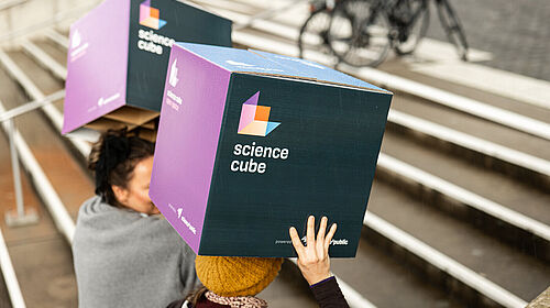 Zwei Papphocker, auf denen "science cube" steht, werden von zwei Personen eine Treppe hochgetragen.