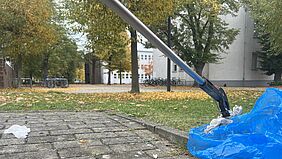 Mit einem Greifer wird ein herumliegendes Taschentuch in einen Müllbeutel gelegt. Im Hintergrund ist der herbstliche Campus zu erkennen.