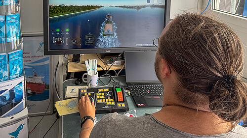 Person mit zusammengebundenem Haar steuert an einem Schiffs-Simulator mit mehreren Kontrollgeräten vor einem Bildschirm. Auf diesem iist eine Wasserstraße mit einem Schiff zu sehen.
