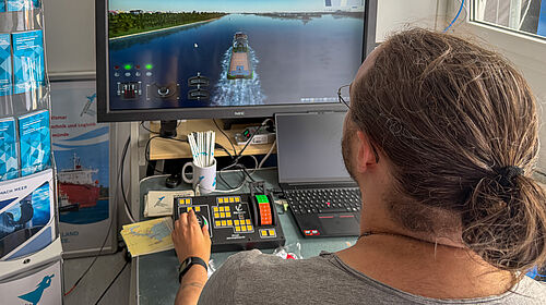 Person mit zusammengebundenem Haar steuert an einem Schiffs-Simulator mit mehreren Kontrollgeräten vor einem Bildschirm. Auf diesem iist eine Wasserstraße mit einem Schiff zu sehen.