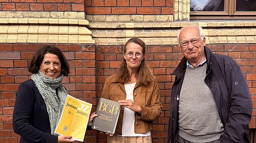 Drei Personen stehen vor einer Backsteinfassade; zwei von ihnen halten juristische Fachpublikationen in den Händen.
