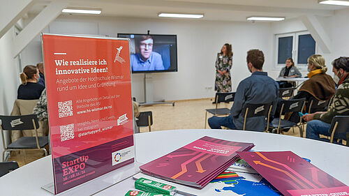 Im Vordergrund sind Infomaterialien zur StartUp-Expo auf einem Tisch  zu sehen während im Hintergrund Teilnehmende in der StatUpYard den Beitrag eies online zugeschalteten Teilnehmers auf einem Bildschirm verfolgen.n n 