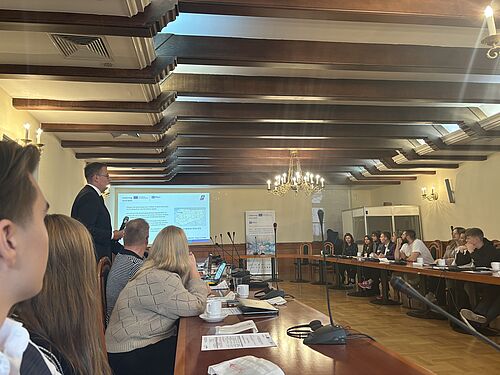 In einem Konferenzraum mit Holzdecke und Kronleuchtern hält ein Mann im Anzug eine Präsentation. Teilnehmende sitzen an einem U-förmigen Tisch, hören zu und haben Mikrofone sowie Unterlagen vor sich. Auf der Leinwand ist eine Folie mit Text und Karte zu sehen. Die Szene wirkt formal und professionell.