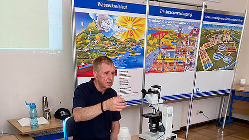 Bertram Jug sitz hinter einem Tisch und schaut in ein Mikroskop. Hinter ihm stehen Schautafen zum Thema Wasserkreislauf.