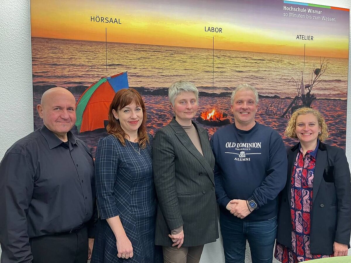 Five people stand side by side, smiling at the camera. In the background is a large beach sunset photo labeled “Lecture Hall,” “Laboratory,” and “Studio,” along with the tagline “Wismar University – 10 minutes to the water.” The individuals are dressed in a mix of formal and casual attire.