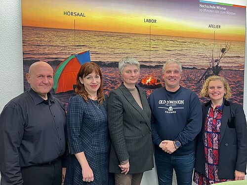 Five people stand side by side, smiling at the camera. In the background is a large beach sunset photo labeled “Lecture Hall,” “Laboratory,” and “Studio,” along with the tagline “Wismar University – 10 minutes to the water.” The individuals are dressed in a mix of formal and casual attire.