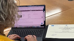 Über die Schulter einer Frau mit blonden kinnlangen Haaren fotografiert ist ein Laptop zu sehen. Sie hat ihre Hände auf dem Touchpad. Neben dem Laptop liegt ein bedrucktes Papier. Darauf sind die Worte Certificate zu lesen.