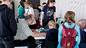 Ein Mann, umringt von Kindern, sitzt an einem Tisch und hilft einem Mädchen bei ihrem Lötprojekt.
