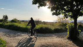 Ein Radfahrer ist vor der utnergehenden Sonne als Silhoutte zu erkennen. Er fährt einen Küstenweg entlang, so dass die Ostsee zu sehen ist.