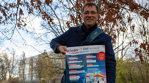 Professor Hillmann steht auf einer Wiese, die mit bunten Blättern zugedeckt ist. In den Händern hält er ein Plakat. Im Hintergrund sind Gebäude der Hochschule zu sehen.
