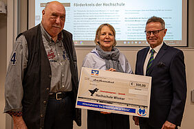 Die Vereinsvorsitzende hält einen ca. einen Meter breiten Scheck in der Hand. Die beiden Herren stehen links und rechts neben ihr. Im Hintergrund ist die Webseite des Förderkreises zu sehen.