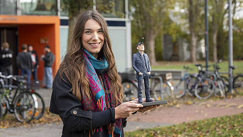 Lousia steht mit der rund 20 cm hohen Preisskulptur cor dem GEbäude der Fakultät Gestaltung. Die Skulpur ist ein Mann in blauer Kleidung. Aus seiner Stirn ragt ein Stift heraus.