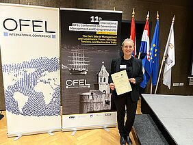 Dr. Laima Gerlitz mit Teilnahmezertifikat der 11. OEFL-Konferenz vor einem Veranstaltungsplakat im Sitzungssaal.