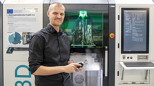 : A man in a black shirt stands in front of a multi-material printer, a large machine resembling a cupboard, with an illuminated interior and a large screen on the right.