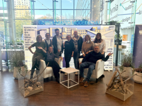 Ten people, including men and women, are standing and sitting in two rows in front of a banner reading “Gdynia Miasto Modernizmu.” Behind them is a large glass wall with a view of Gdynia’s city centre.