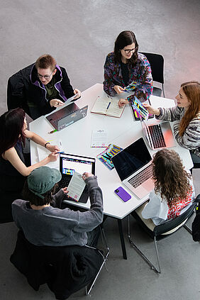 Sechs Personen sitzen im Kreis an einem sechseckigen Tisch. Auf ihren Laptops sind Grafik-Entwürfe zu sehen.