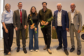 Sechs Personen stehen nebeneinander vor einer weißen Wand. Zwei in der Mitte halten jeweils eine Auszeichnung und Blumen. Alle lächeln.