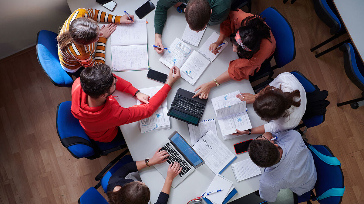 Introduction_Image_Symbolbild_WR_Master_SL_2023 Introduction_Image Studiengang Wirtschaftsrecht Master Studierende sitzen um einen Tisch herum auf dem Unterlagen verstreut sind, Draufsicht