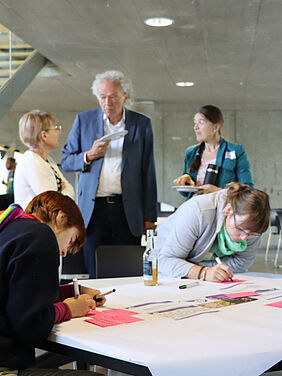 ZWi Frauen sitzen an einem Tisch und schreiben etwas auf klene farbige Zettel. Dahnter steht ein Mann, der mit zwei weiteren Frauen spricht.