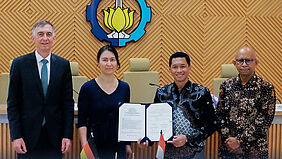 Die vier PErsonen stehen vor einer mit Holz getäfelten Wand, an der ein großes Logo der ITS hängt. Der REktor der ITS hält eine geöffnete Mappe in die Kamera. Darin befindet sich die unterzeichnete Urkunde.