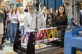 Man sieht eine bunte Brücke aus Papier, die in eine MEtallvorrichtung eingespannt ist. Dahinter sind unscharf Schülerinnen zu erkennen, die zuschauen.