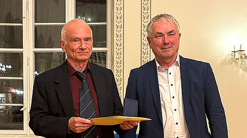 Die beiden Personen stehen nebeneinander und es sind deren Oberkörper zu sehen. Der Ausgezeichnete hält eine Mappe in der Hand.