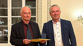 Die beiden Personen stehen nebeneinander und es sind deren Oberkörper zu sehen. Der Ausgezeichnete hält eine Mappe in der Hand.