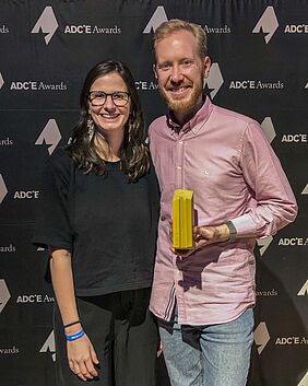 Die beiden Personen stehen nebeneinander vor einer schwarzen Leinwand, auf der das Logo des ADCE zu sehen ist.