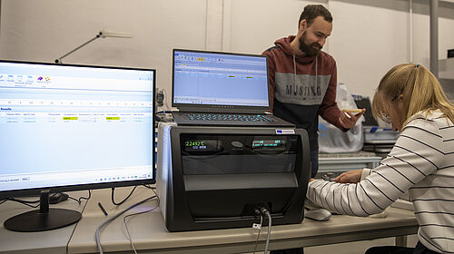 Zwei Personen an einem Tisch mit einem Messgerät, an dem Hot Disk TPS 2500 S steht, und zwei Bildschirmen. Eine männliche Person steht mit einem kleinen Gegenstand in der Hand. Die andere, weibliche Person sitzt und arbeitet am Gerät.