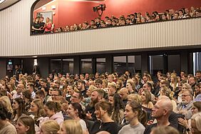 Publikum in einem Theater bei einem Festakt, sitzend und stehend auf zwei Ebenen. Sie blicken aufmerksam nach vornund manche klatschen.