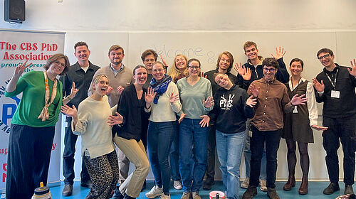 15 Personen stehen in zwei Reihen nebeneinander und winkend fröhlich lachend in die Kamera. Links im Hintergrund ist ein Roll-up zu sehen, auf dem der Name der Doktorandenvereinigung steht.