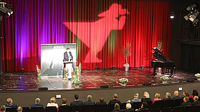 Mit Blick von der Empore ist der Minister als Redner auf der Theaterbühne zu sehen sowie einige Gäste, die in der ersten und zweiten Reihe sitzen.