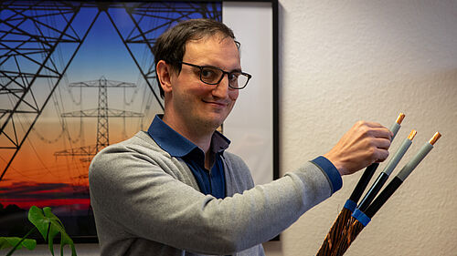 Der PRofessor schaut seitwärts in die Kamera. Er hält ein ca. 1 Meter langes Kabel in der Hand. Das ist aufgeschnitten und besteht aus drei kleineren Kabeln.