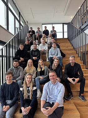 Group photo at the Pauli House About 25 people are sitting on a wide wooden staircase in a light-filled room, smiling at the camera.