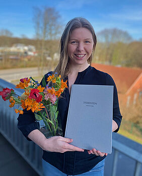 Auf dem Bild ist Dr. Johanna Ender mit einem Blumenstrauß und ihrer gedruckten Doktorarbeit zu sehen.