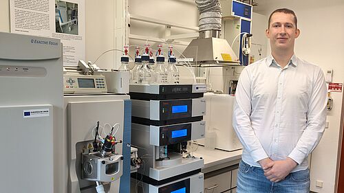 Der junge Mann trägt ein weißes Hemd und eine Jeans. Er steht in einem Labor neben Apparaturen. Im Hintergrund sind weitere Labormöbel und technische Geräte zu sehen.