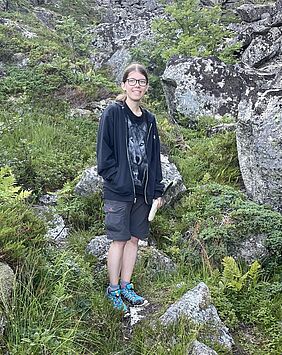 Die Person steht in Wanderkleidung an einem Hangzwischen Felsblöcken und kleinen Pflanzen wie Moose, Farne und Sträucher.