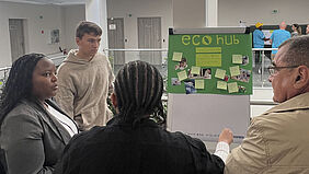 Vier Personen stehen vor einem grünen Plakat mit der Überschrift 'eco hub' und mehreren angehefteten Zetteln in einem hellen Raum.