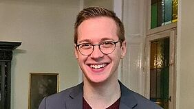 Junger Mann im Anzug und mit Brille lacht freundlich, im Hintergrund ein Raum mit bunten Fenstern, Bild und schönem alten Schrank..