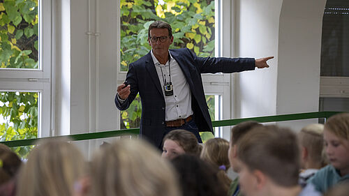 Der MAnn ist über die Köpfe einiger Kinder hinweg zu sehen. Er steht am Rande des Hörsaals und zeigt auf ein FEnster. 