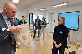 Der Prorektor hält den Prototypen in der Hand, ein Pflanzgefäß aus Ton, und stellt der Studentin eine Frage.