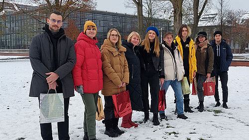 Es sind acht ukrainische Studierende mit kleinen Weihnachtsgeschenken in der Hand gemeinsam mit der Leiterin des International Office im Schnee stehend vor der Bibliothek zu sehen.