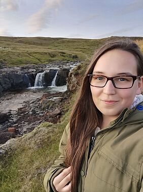 Die junge Frau steht mitten in iner Graslandschaft vor einem Bach. Sie ist im Porträt zu sehen.