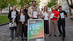 Die sieben Personen stehen nebeneinander. Im Vordergrund ist in einem Aufsteller das Plakat für den Science Slam zu sehen. Die beiden GEwinner halten die Siegerkrone über ihrem Kopf.