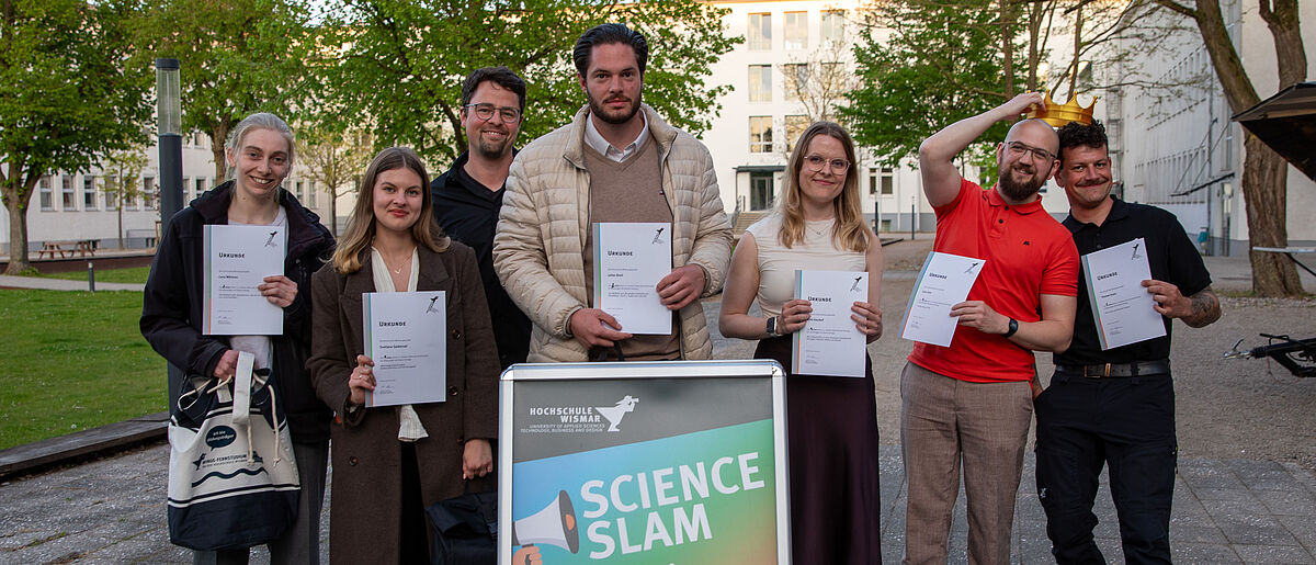 Die sieben Personen stehen nebeneinander. Im Vordergrund ist in einem Aufsteller das Plakat für den Science Slam zu sehen. Die beiden GEwinner halten die Siegerkrone über ihrem Kopf.