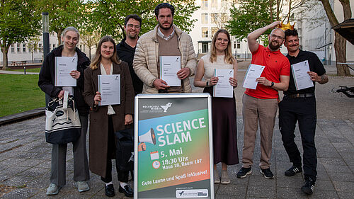 Die sieben Personen stehen nebeneinander. Im Vordergrund ist in einem Aufsteller das Plakat für den Science Slam zu sehen. Die beiden GEwinner halten die Siegerkrone über ihrem Kopf.