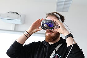 A young man with a beard is putting on an Apple Vision Pro headset. He is wearing a black hoodie and wristbands. The bright seminar room includes a ceiling light and air conditioning unit in the background.