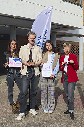 Die vier Personen stehen nebeneinander und halten eine Urkunde, einen kleinen gehäkelten Fisch und einen Gutschein in der Hand. Hinter ihnen an der Wand ist eine BEachflag mit der Aufschrift "Medienkompetenzpreis" zu sehen.